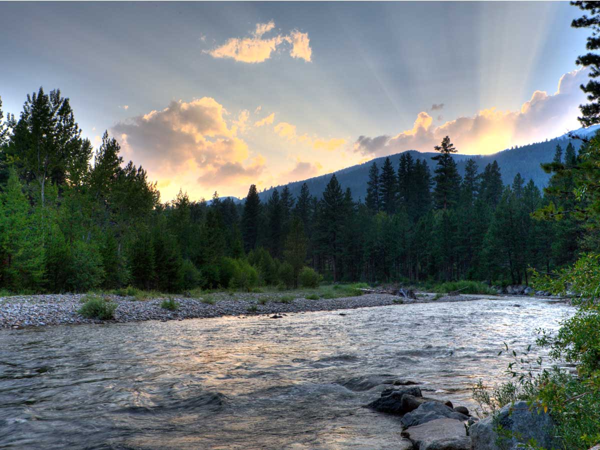 Image of sun setting behind mountain with forested stream in foreground.